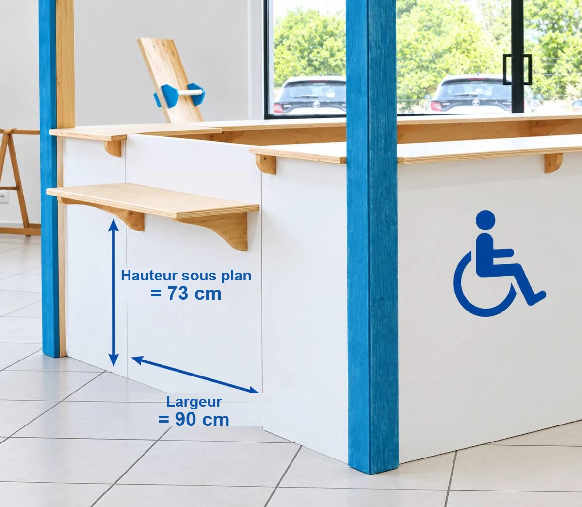 Comptoir blanc et bleu équipé d'une tablette rabattable en bois, coté hauteur sous plan 73 cm et largeur 90 cm, à côté du pictogramme d'accessibilité